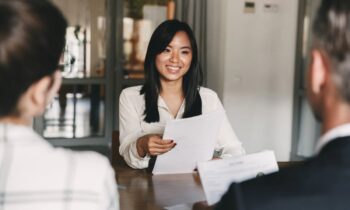 A woman sitting down in an office across from two people for an interview. She's handing them her resume.