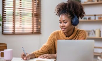 College student working from home, using a laptop, wearing headphones, and writing in a notebook for blog post side hustles for college students.