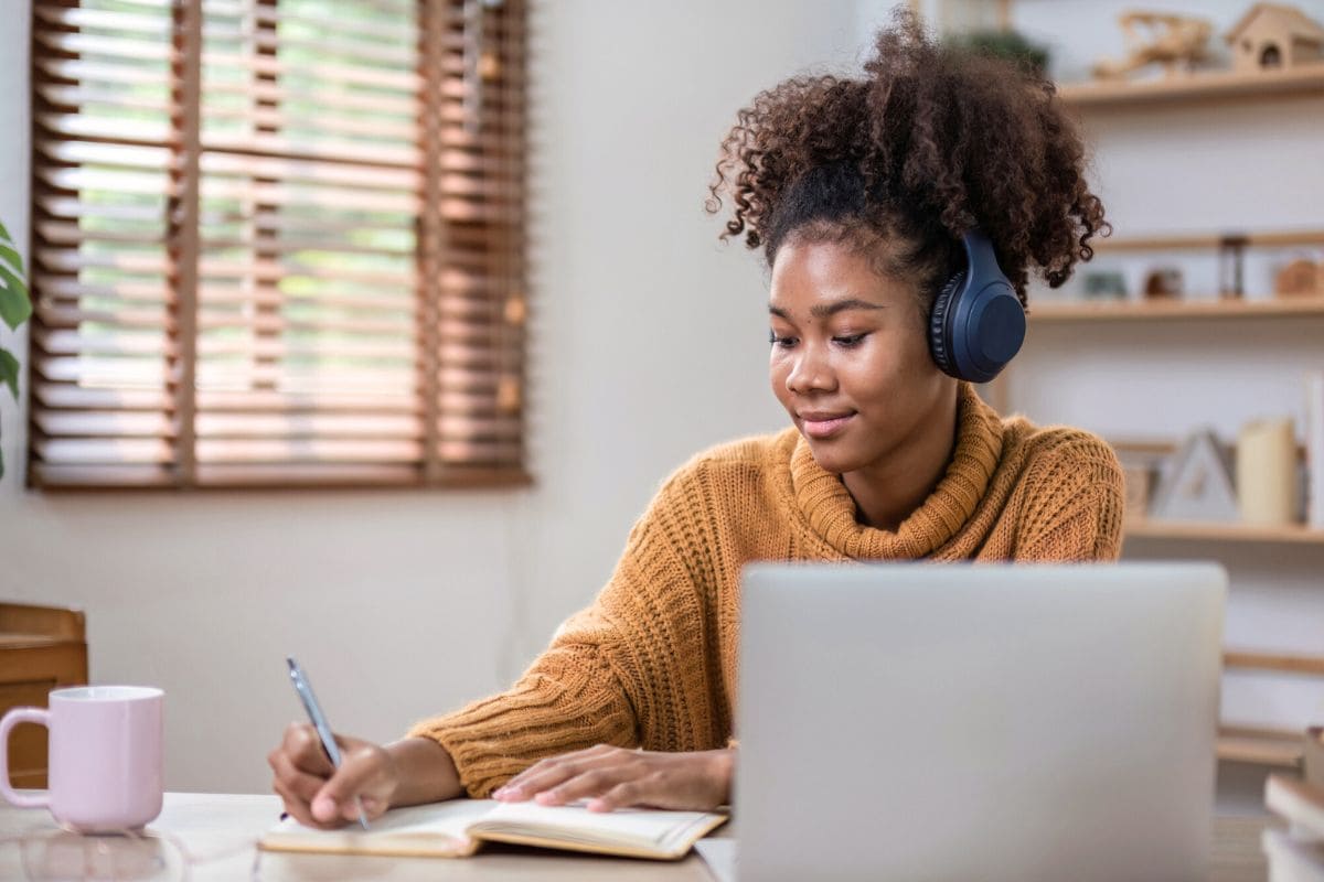 College student working from home, using a laptop, wearing headphones, and writing in a notebook for blog post side hustles for college students.