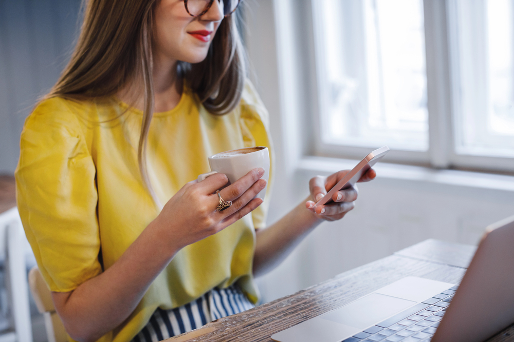 A woman wearing glasses drinking coffee earning free gift cards on her phone