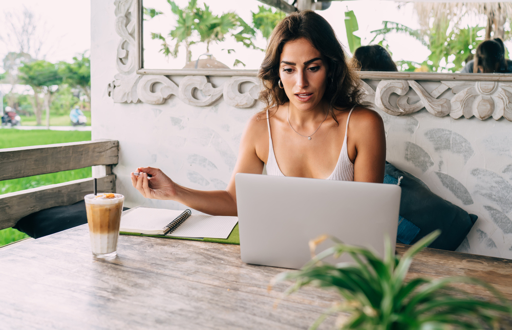 A young woman working at an outdoor cafe reading an article online titled how to make extra money online