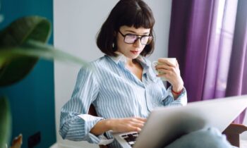 A woman working from home on a laptop and holding a cup of coffee trying to find out someone's salary