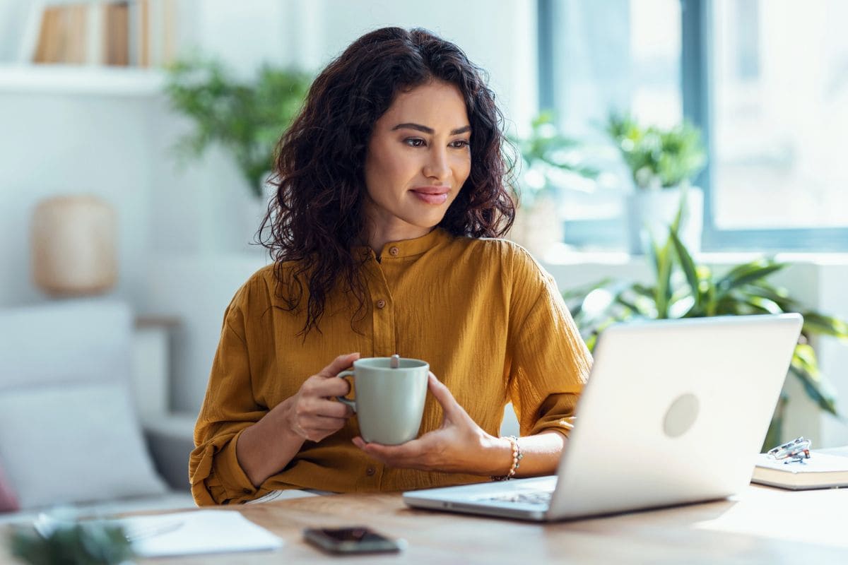 A woman working from her home office desk, holding a coffee cup, smiling, and reading on her laptop for blog post get promoted at work.