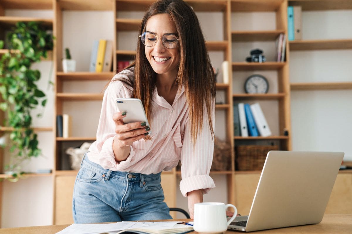A work at home business woman standing at a desk in her home office, looking at something on her phone for blog posy make money on Pinterest