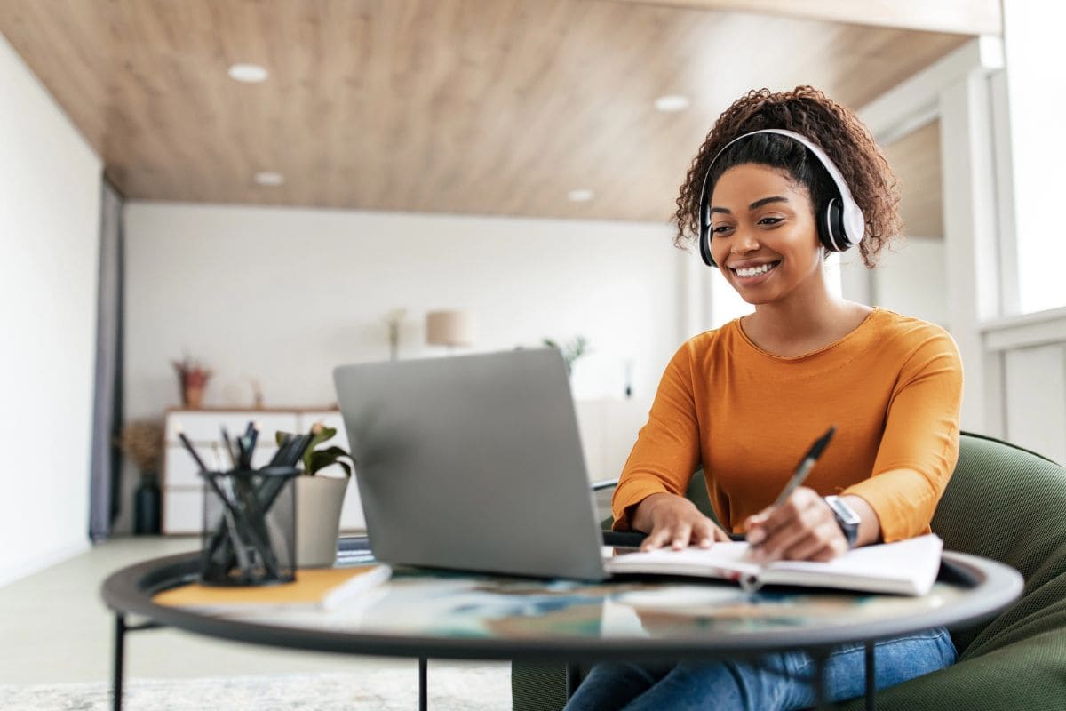 A freelancer working remotely from her kitchen table, using a laptop, wearing headphones, and writing in a notebook for blog post sell services online
